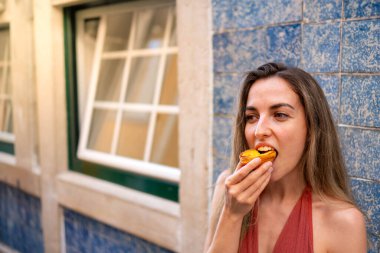 Yakın plan, genç bir kadının geleneksel Portekiz Pastel de Nata 'sının tadını çıkarırken cepheden görünüşü, Gün ışığında yatay olarak yakalanıyor.