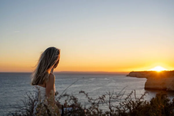 Young Woman in Profile, wearing White Dress, Stands on a Cliff Overlooking the Ocean at Sunset,