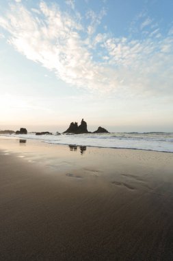 Altın saatte sakin bir sahil, kaya, su, kum ve gökyüzünün harmoni içinde birleştiği, dikey bir çerçevede yakalandığı