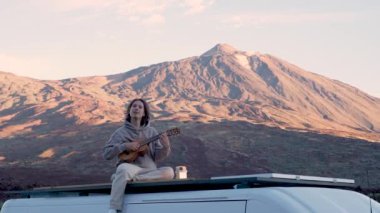 Arka planda dağ olan bir minibüsün tepesinde otururken gitar çalma becerilerini sergileyen uzun saçlı genç bir adamın inanılmaz yeteneğine tanık olun.