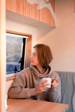 Moment of tranquility as a young man enjoys a warm beverage inside the cozy wooden interior of his self-converted van, showcasing a DIY travel lifestyle. clipart