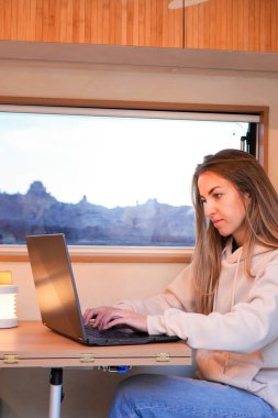 A digital nomad is focused on her laptop while working from the well-lit interior of her van, with the scenery of dusk visible through the window. clipart