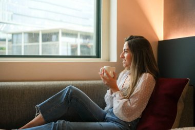 Genç bir kadın rahatça bir otel odasının penceresinde oturur, dışarı bakarken sıcak bir fincan kahvenin tadını çıkarır, rahatlamış bir sabahın basit zevkini ön plana çıkarır..