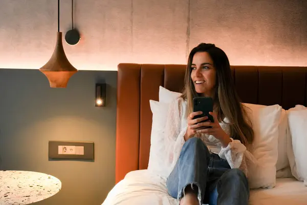 Happy young woman guest sitting on a bed in a luxury hotel room, engaged with her cell phone.