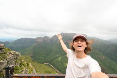Neşeli genç bir adam arka planda nefes kesici Drakensberg sıradağları olan bir selfie çekiyor. Beyaz bir tişört ve kep giyerek, geniş yeşil bir manzara arkasında bulutlu bir gökyüzünün altında uzanan bir bakış açısının üzerinde duruyor..