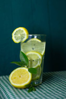 Güzel bir arka planda taze limon ve yeşil yapraklarla süslenmiş şeffaf bir limonata bardağı.