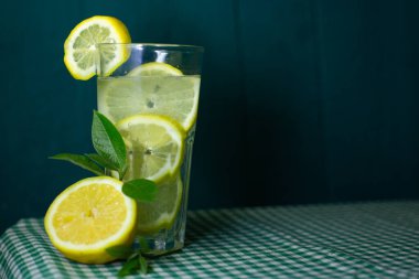 Güzel bir arka planda taze limon ve yeşil yapraklarla süslenmiş şeffaf bir limonata bardağı.