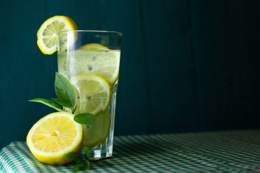 Güzel bir arka planda taze limon ve yeşil yapraklarla süslenmiş şeffaf bir limonata bardağı.