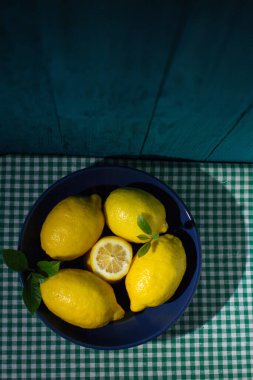 Koyu mavi bir kasede yaprakları olan taze limonlar kareli masa örtüsünün üzerinde.