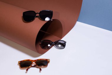 Kahverengi tasarım kağıdının yanında beyaz ve mavi bir zemin üzerinde geniş çerçeveli gözlük.