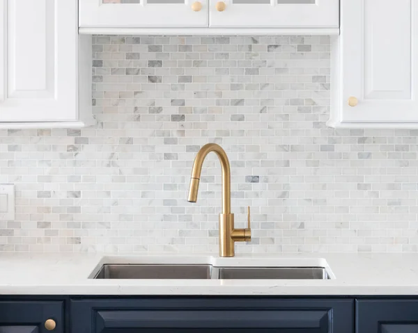 A kitchen sink detail shot in a white and blue kitchen with a gold faucet, marble countertop, and small marble subway tile backsplash.
