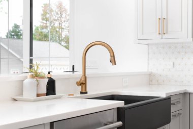 Altın musluğu, siyah çiftlik evi lavabosu, beyaz graniti ve fayanslı döşemesi olan yenilenmiş modern bir çiftlik evi mutfağında güzel bir lavabo. Etiket yok.