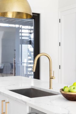 A gold kitchen faucet in a white kitchen with a marble countertop, gold light fixture hanging down, and a bowl of green apples.