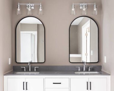 A bathroom with a white vanity and black hardware, chrome faucets on a quartz countertop, arched mirrors, and brown walls.