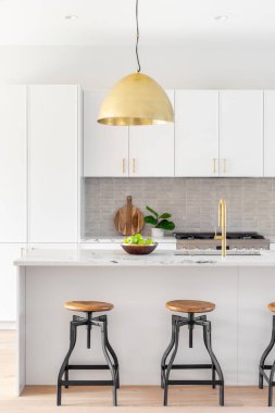 A kitchen detail with white cabinets, gold faucet and light hanging over the island with bar stools, and a tiled backsplash.