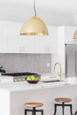 A kitchen detail with white cabinets, gold faucet and light hanging over the island with bar stools, and a tiled backsplash.