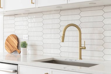 A beautiful kitchen faucet detail with white cabinets, a gold faucet, white marble countertops, and a brown picket ceramic tile backsplash.