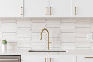 A beautiful kitchen faucet detail with white cabinets, a gold faucet, white marble countertops, and a brown picket ceramic tile backsplash.