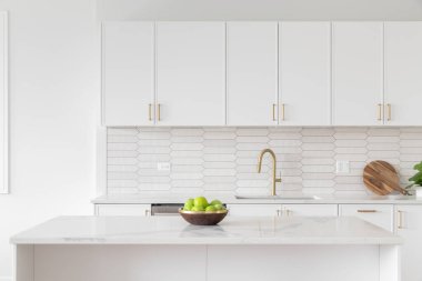 A beautiful kitchen faucet detail with white cabinets, a gold faucet, white marble countertops, and a brown picket ceramic tile backsplash.
