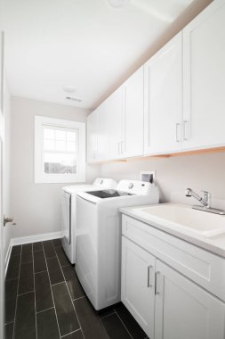 A laundry room with dark grey tiled floor, white cabinets and appliances, and a sink with a chrome faucet.