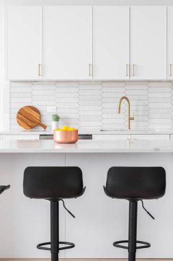 A kitchen detail with white cabinets, a picket tiled backsplash, gold faucet and hardware, and bar stools sitting at the marble island.