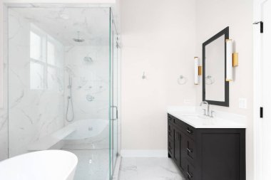 A bathroom with a dark grey cabinet, gold sconce lights around the mirror, and a walk-in shower with marble tiles.