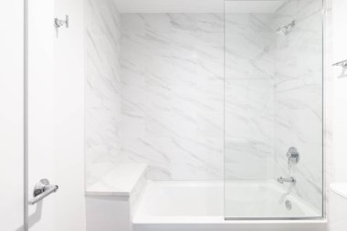 A shower with white marble tile and glass walls, a marble hexagon tiled floor, bench seat, and chrome showerhead.