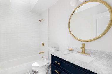 A white bathroom with a blue vanity cabinet, granite countertop, pattern tile flooring, and gold fixtures and mirror.