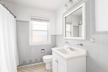 Beyaz dolabı, gri fayanslı duvarları ve kahverengi döşemesi olan bir banyo.. 
