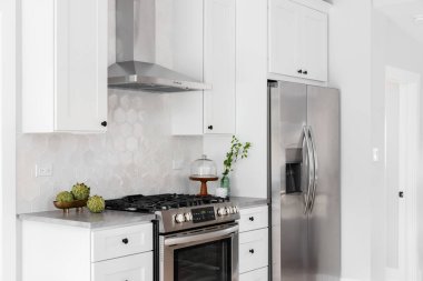 A kitchen detail with white cabinets, stainless steel appliances, tan hexagon tile backsplash, and decorations on the grey marble countertop. clipart