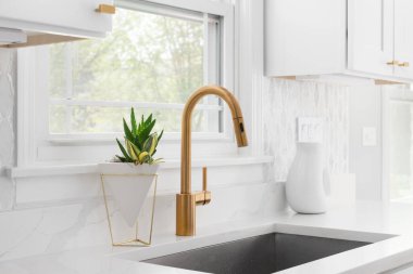 A kitchen sink detail with a gold faucet, marble tile backsplash, and white cabinets. clipart