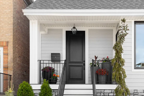A black front door on a suburban grey and white house with plants on a small front porch and black railing.