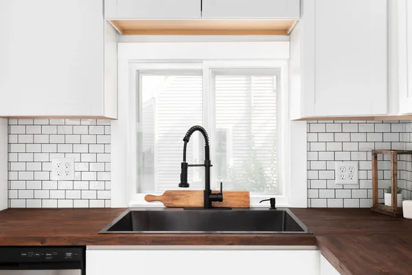 A black kitchen sink and faucet detail in a white kitchen with a butcher block countertop, white subway tile backsplash, and cozy decor.