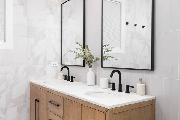 A bathroom with marble and stacked vertical subway tiles, a white oak vanity cabinet, black framed square mirrors and faucets, and marble countertop.