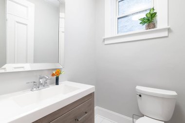 Küçük bir banyo, ahşap bir dolap, beyaz lavabo ve gri duvarlar..