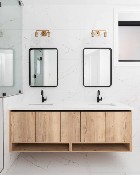 A bathroom detail with a floating wood cabinet, white marble tiled wall and floor, and a gold light fixture mounted above the black frame mirror.