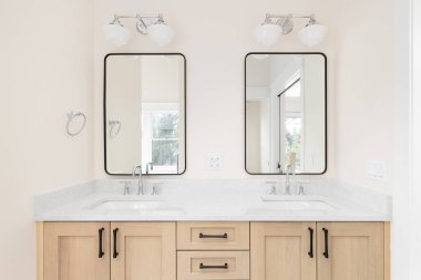 Beyaz meşe rengi makyaj dolabı, mermer tezgah, krom musluklar ve siyah çerçeveli aynaların üzerinde asılı ışıklar olan rahat bir banyo detayı..