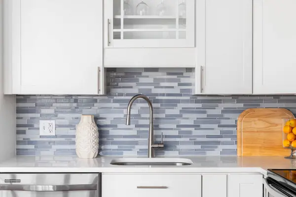 A kitchen faucet detail with a blue glass tile backsplash, white cabinets, decorations on the marble countertop, and a stainless faucet.