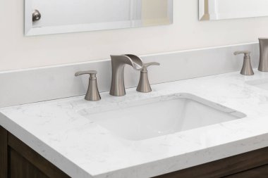 Bronz, geniş bir musluğu, beyaz mermer tezgahı ve koyu ahşap bir dolabı olan bir banyo musluğu..