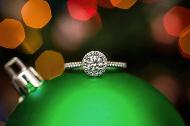 Arkasında bulanık ışıklar olan yeşil bir Noel süsü takan nişan yüzüğü..
