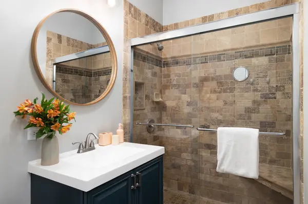 A bathroom with a blue cabinet wooden circular mirror above a white sink, and a shower with brown stone tiles.