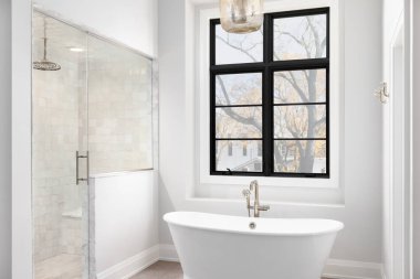 A bathroom detail with light hanging above a freestanding soaker tub, a black large framed window, and a tiled walk-in shower. clipart