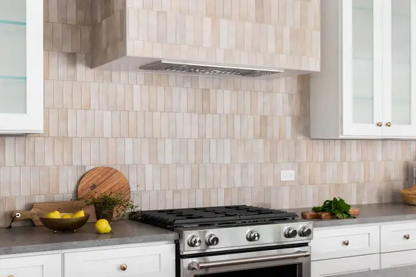A kitchen oven and hood detail with brown rectangle tiles, stainless steel oven, white cabinets, and cozy decor on grey stone countertops.