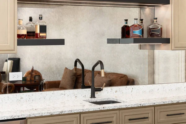 A wet bar with brown cabinets, a white countertop, bottles sitting on floating shelves, and a black and gold faucet. No brands or labels.