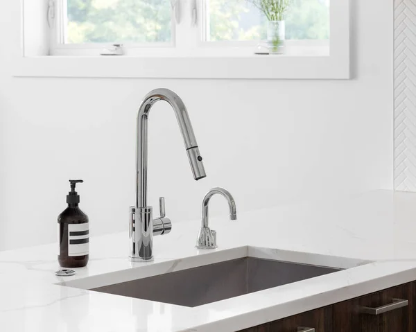 A faucet detail in a modern kitchen  on a white marble countertop, wood cabinet, and chrome faucet.