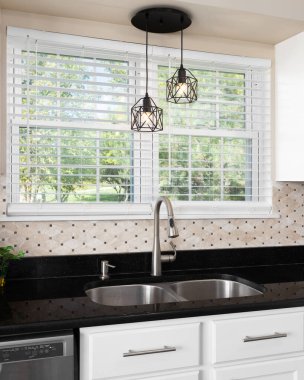 Siyah kuvars tezgahı olan bir mutfak lavabosu ayrıntısı, kahverengi taşlı bir zemin kaplaması, ve parlak paslanmaz çelik musluğun üzerinde asılı ışıklar..