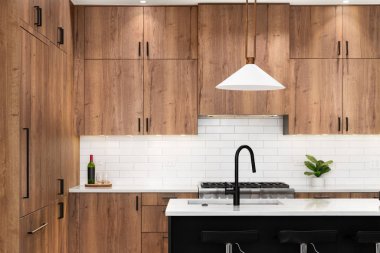 Ahşap dolaplı bir mutfak müfrezesi, siyah bir musluk, metro döşemesi, ve siyah adanın üzerinde asılı altın ışıklı eşyalar ve mermer tezgahlar..