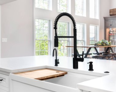 Beyaz bir mutfakta beyaz bir çiftlik evi lavabosu ve krom lambası mermer tezgahın üzerinde asılı duran siyah bir musluk..