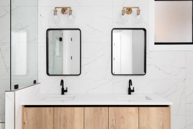 Ahşap bir dolabı, beyaz mermer döşeli bir duvarı ve siyah çerçeveli aynanın üzerine monte edilmiş altın bir aydınlatma armatürü olan bir banyo ayrıntısı..