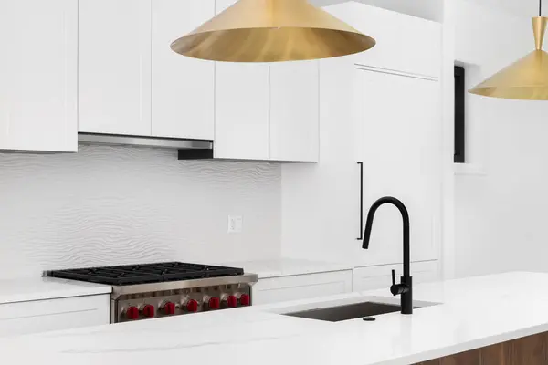 A kitchen detail with a gold light fixtures hanging over a white marble island and black faucet, white modern cabinets, stainless steel stove, and a wavy tiled backsplash.